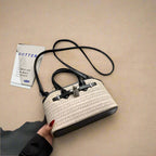 Handbag with textured design held by a hand on a white surface with a gray garment and book in the background.